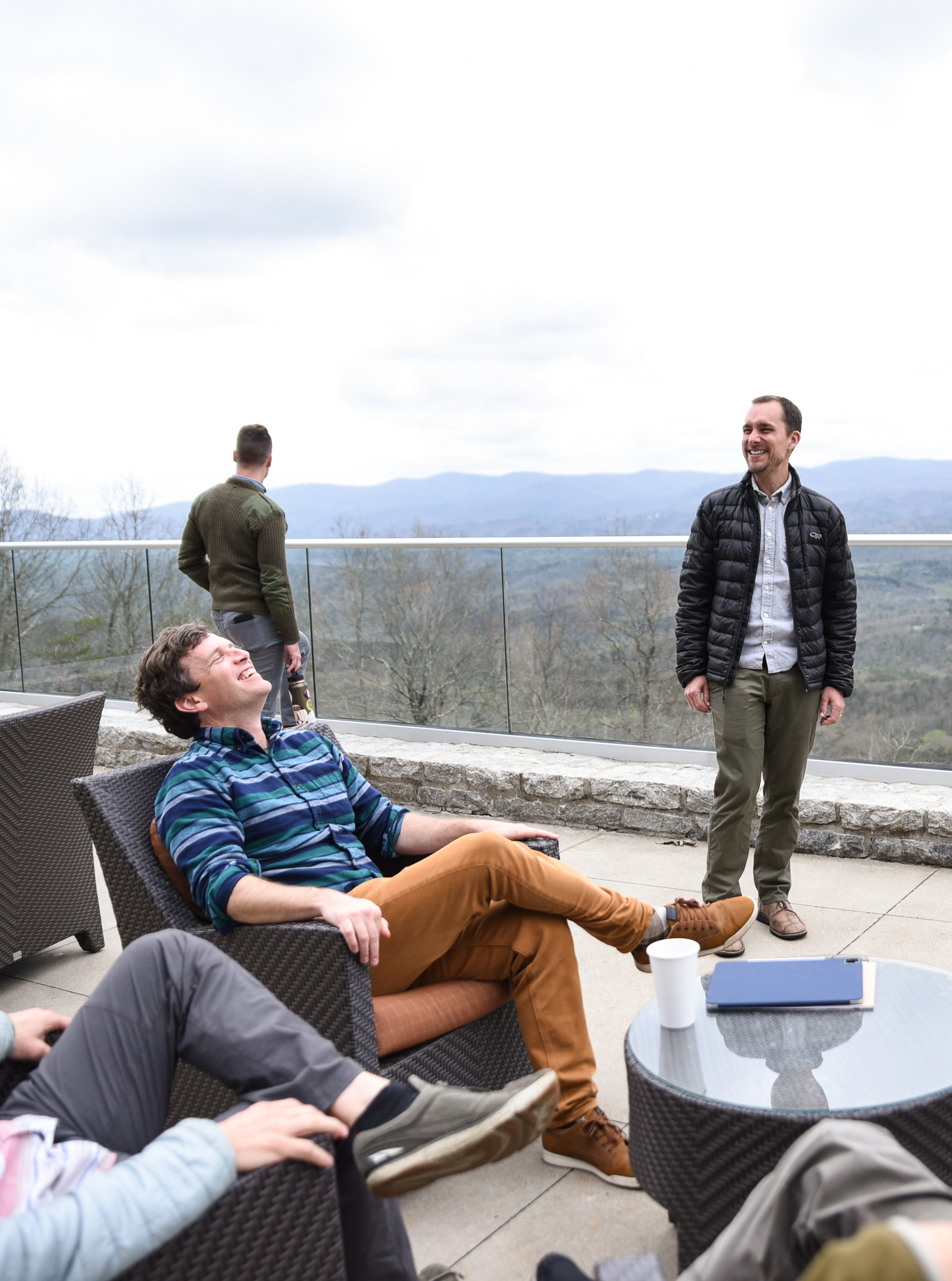 Two men talk to each other on a patio overlooking the mountains. The one on the left is wearing a blue striped pull over and rusty orange pants. His head is thrown back, laughing.