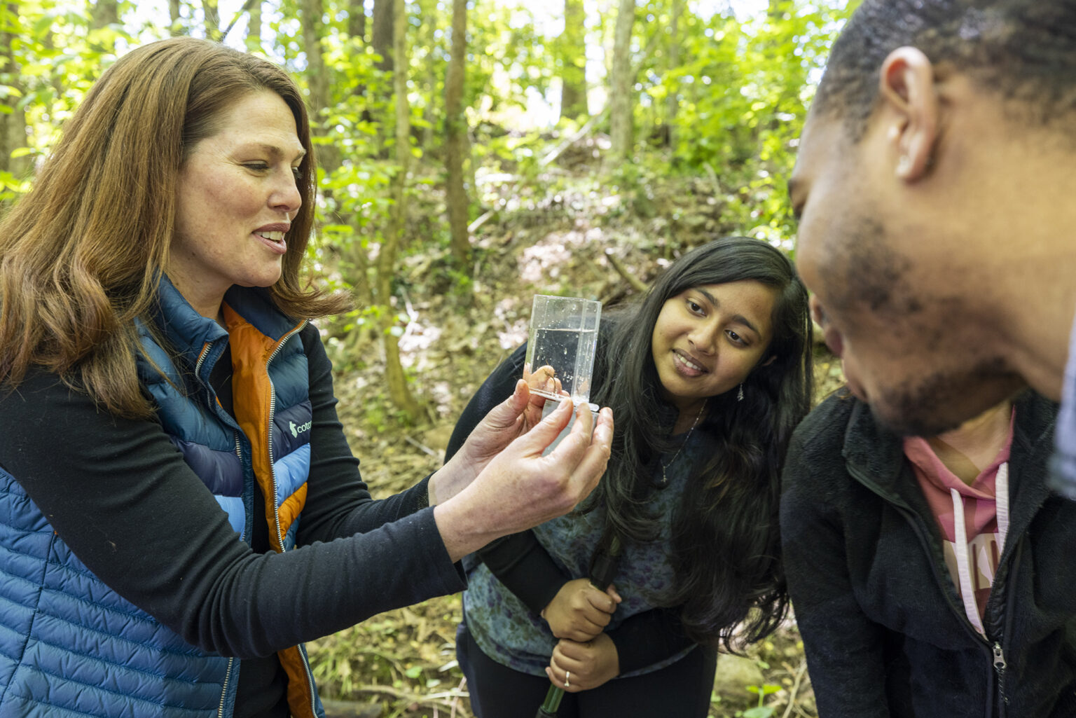 A woman wearing a blue and orange puffy vest with a black shirt smiles as she looks at a small glass box containing an aquatic specimen. Three students gather around to look closely at it. In the background, sunlight filters through green tree canopy.