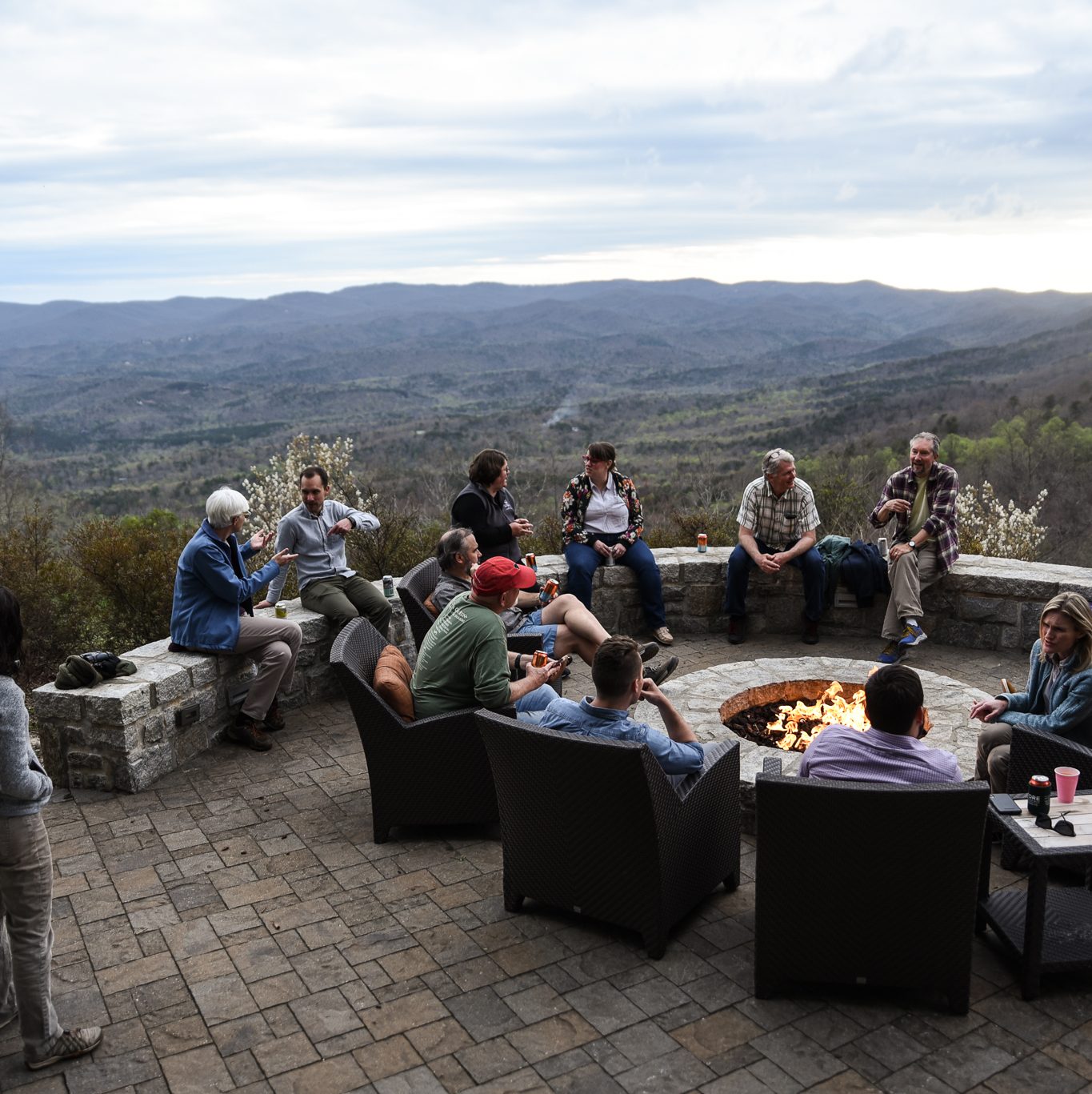 About 15 people sit around a fire, some in chairs and others on a stone wall. Behind them, the sun sets on the mountains